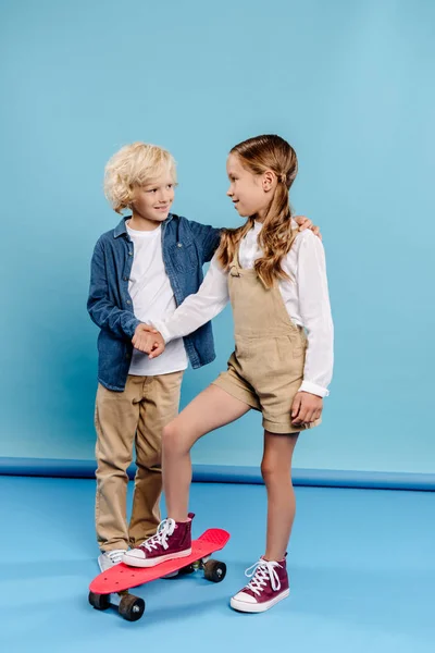 Lächelnd und niedlich Junge umarmt Freund auf Penny Board auf blauem Hintergrund — Stockfoto