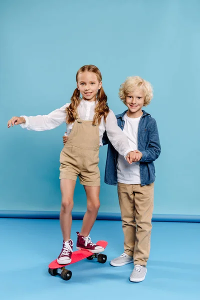 Lächelnder und netter Junge, der die Hand eines Freundes auf einem Penny-Board auf blauem Hintergrund hält — Stockfoto