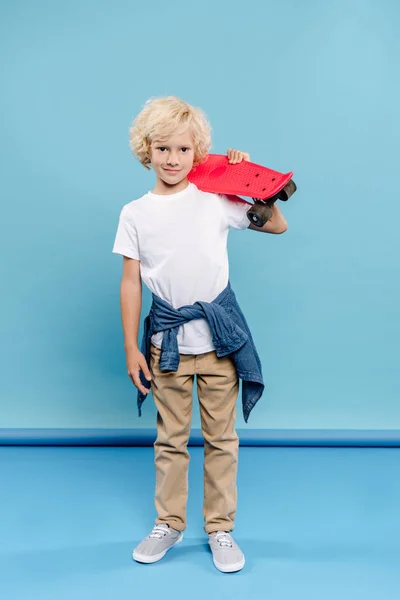 Lächelndes und nettes Kind, das in die Kamera schaut und Penny-Board auf blauem Hintergrund hält — Stockfoto