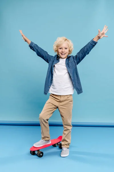 Enfant souriant avec les mains tendues debout sur la planche de penny sur fond bleu — Photo de stock