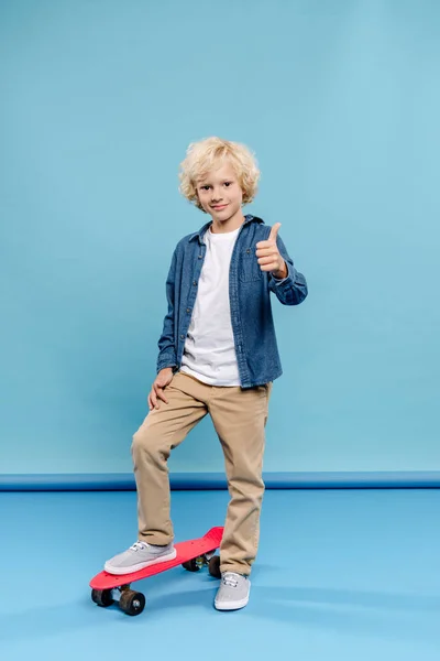 Lächelndes und nettes Kind zeigt wie und steht auf Penny-Board auf blauem Hintergrund — Stockfoto