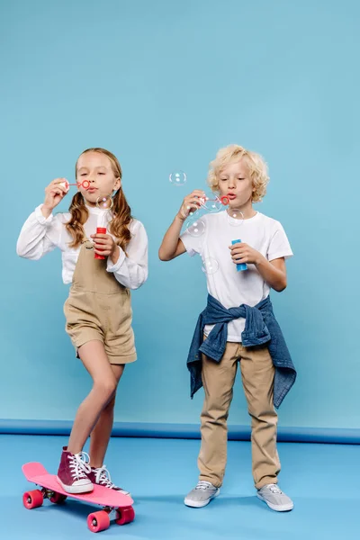Mignons enfants debout sur des planches de penny et soufflant des bulles de savon sur fond bleu — Photo de stock