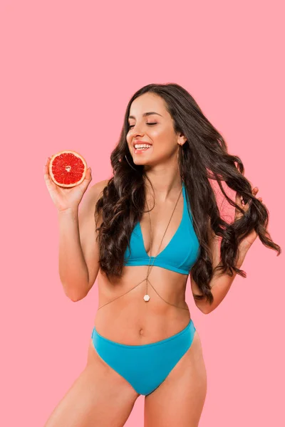 Щаслива жінка в купальнику тримає половину грейпфрута і торкається волосся ізольовано на рожевому — стокове фото