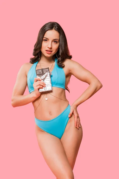 Sexy girl in swimsuit standing with hand in hip and holding sweet chocolate bar isolated on pink — Stock Photo