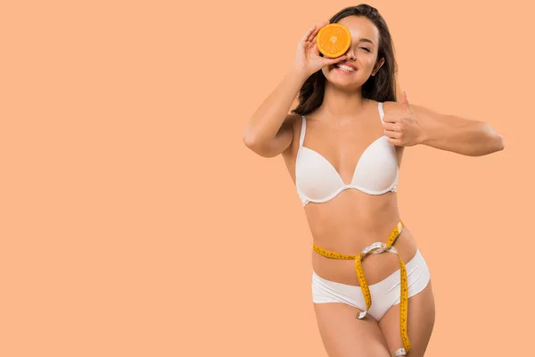 Mujer alegre en ropa interior blanca sosteniendo naranja y mostrando el pulgar hacia arriba aislado en beige - foto de stock