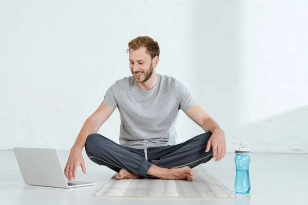 Homme souriant assis dans la pose facile près de l'ordinateur portable et bouteille de sport — Photo de stock