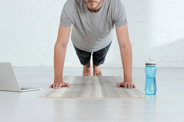 Vista parcial del hombre practicando la postura de la tabla del antebrazo cerca de la computadora portátil y la botella de deportes - foto de stock