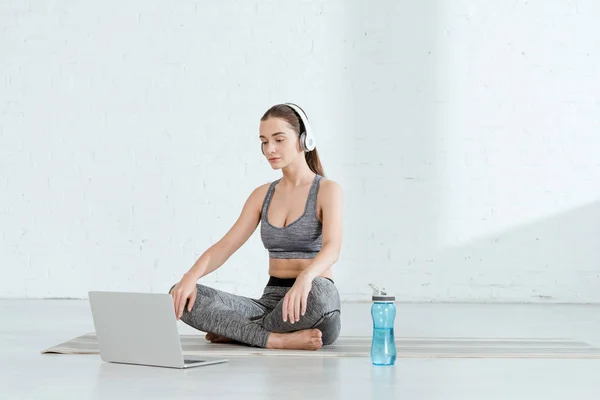 Молода жінка в навушниках, сидячи в легкій позі біля ноутбука і спортивної пляшки — стокове фото