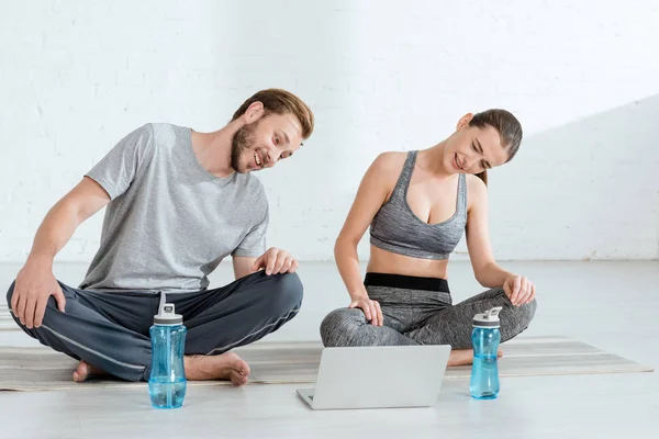 Sonriente hombre y mujer mirando a la computadora portátil mientras está sentado en poses fáciles cerca de botellas deportivas - foto de stock