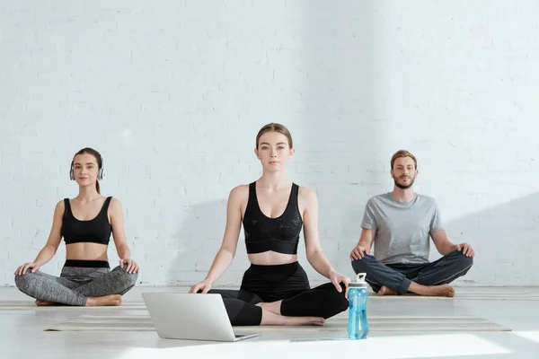 Hombres y mujeres jóvenes practicando yoga en pose de medio loto cerca de laptop y botella deportiva - foto de stock