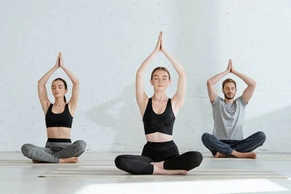 Hombres y mujeres jóvenes practicando yoga en pose de medio loto con las manos levantadas de oración - foto de stock
