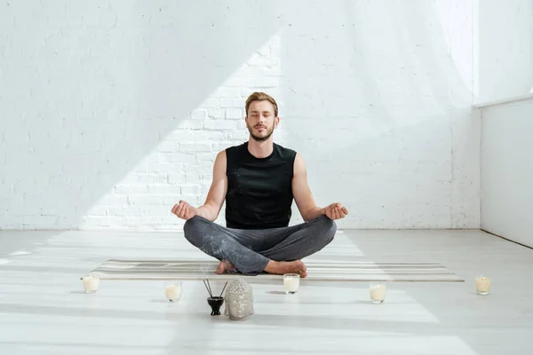 Joven guapo practicando yoga en pose de medio loto cerca de la cabeza decorativa de buda, palos aromáticos y velas - foto de stock