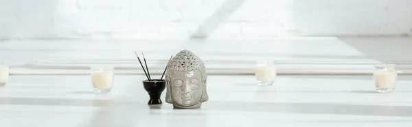Panoramic shot of decorative buddha head near aromatic sticks and candles on white floor — Stock Photo