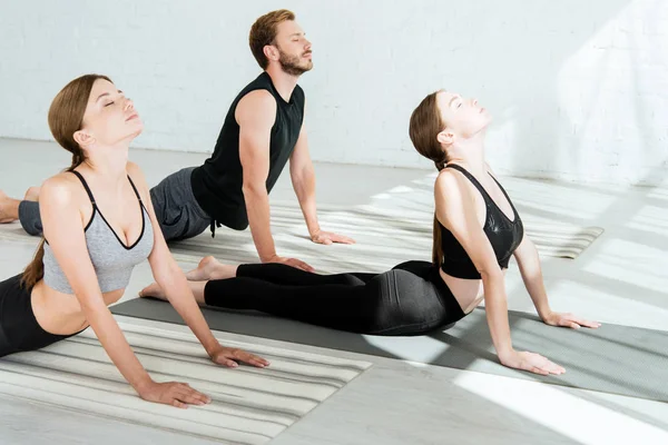 Дві молоді жінки і чоловік практикує йогу у високій позі кобри — стокове фото