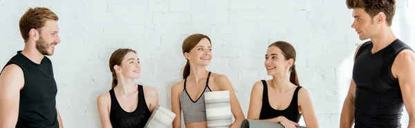 Plano panorámico de mujeres sonrientes con esteras de yoga de pie cerca de hombres guapos - foto de stock