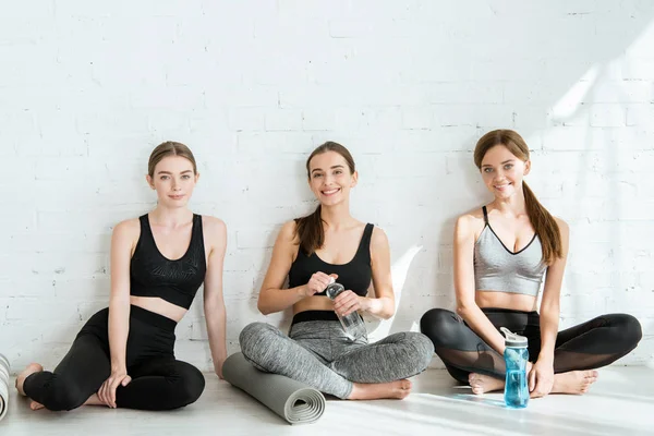 Tres mujeres sonrientes sentadas cerca de la pared en pose fácil con botellas de agua y colchonetas de yoga - foto de stock