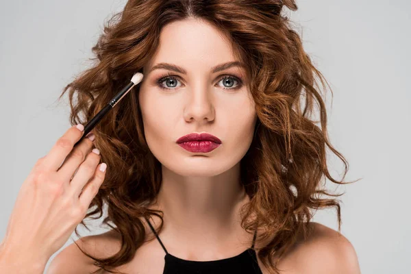 Cropped view of makeup artist apply eye shadow on young curly woman isolated on grey — Stock Photo