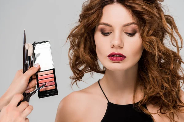 Cropped view of makeup artist holding lip gloss near beautiful woman isolated on grey — Stock Photo