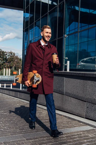 Hombre guapo sosteniendo taza de papel y tablero de penique mientras camina por la calle - foto de stock