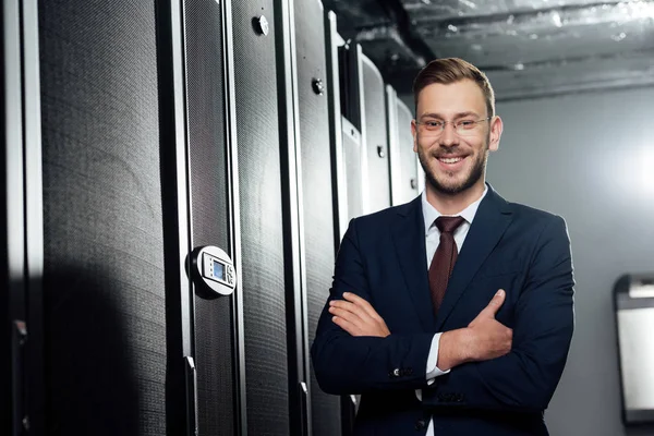 Selective focus of cheerful businessman in suit and glasses standing with crossed arms in data center — Stock Photo