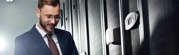 Panoramic shot of cheerful businessman in suit and glasses standing in data center — Stock Photo