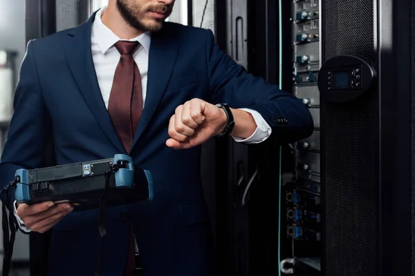 Cropped view of businessman standing with reflectometer and looking at watch in server room — Stock Photo