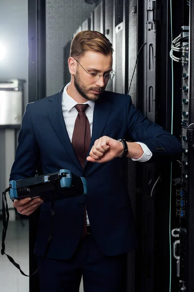 Handsome businessman standing with reflectometer and looking at watch in server room — Stock Photo