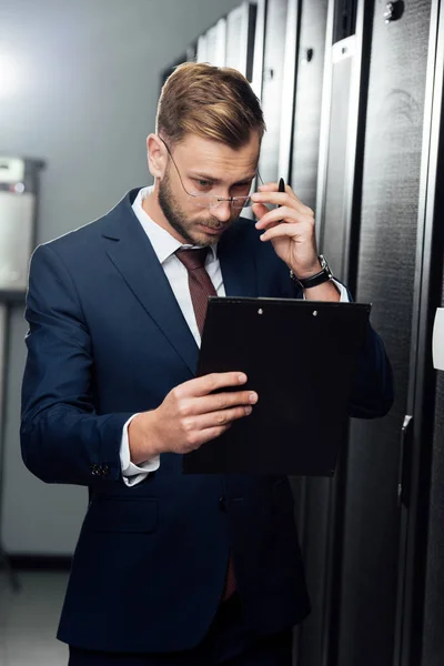 Handsome businessman touching glasses while looking at clipboard — Stock Photo