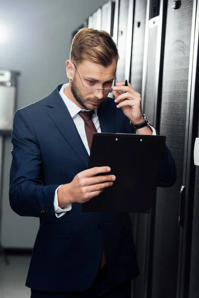 Guapo hombre de negocios tocando gafas mientras mira portapapeles - foto de stock