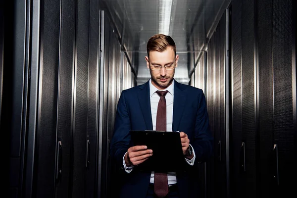 Schöner Geschäftsmann mit Brille blickt auf Klemmbrett in der Nähe von Serverschränken — Stockfoto