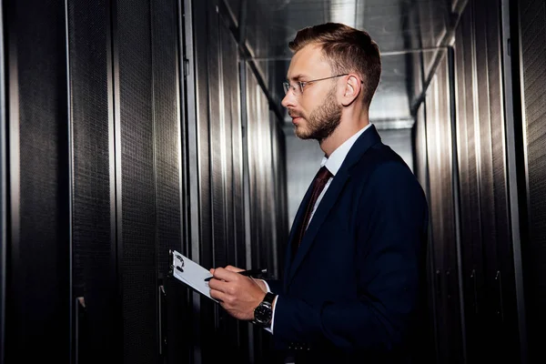 Side view of businessman in glasses holding clipboard near server racks — Stock Photo