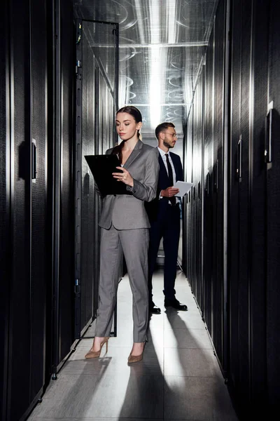 Selective focus of attractive businesswoman holding clipboard near bearded businessman in data canter — Stock Photo