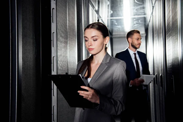 Selective focus of attractive businesswoman holding clipboard near businessman in data canter — Stock Photo