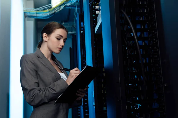 Attraktive Geschäftsfrau schreibt, während sie Klemmbrett im Serverraum hält — Stockfoto