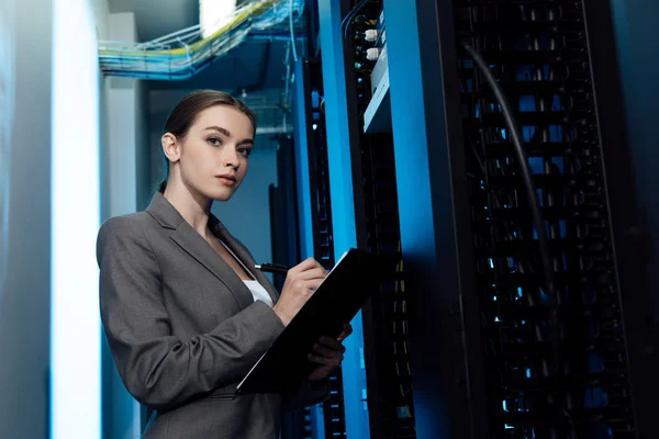 Beautiful businesswoman writing while holding clipboard in server room — Stock Photo