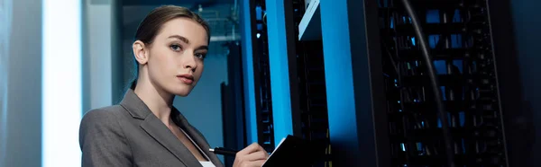 Panoramic shot of beautiful businesswoman writing while holding clipboard in server room — Stock Photo