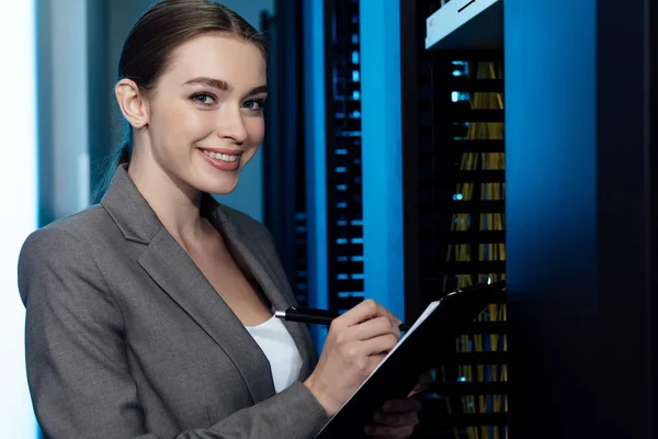 Cheerful businesswoman writing while holding clipboard in server room — Stock Photo