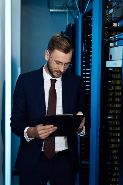 Bel homme d'affaires dans les lunettes tenant presse-papiers dans le centre de données — Stock Photo