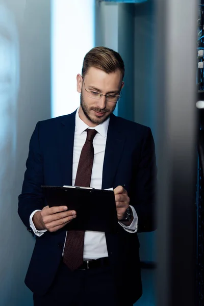Bearded businessman in glasses holding pen and clipboard in data center — Stock Photo