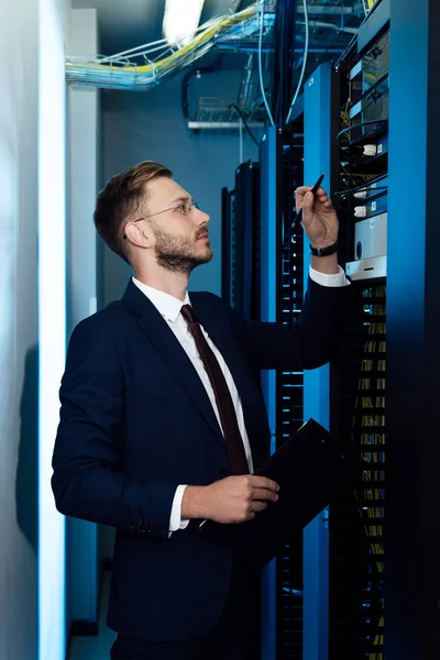 Profil eines gutaussehenden Geschäftsmannes mit Brille, der Stift und Klemmbrett hält, während er auf das Servergestell blickt — Stockfoto