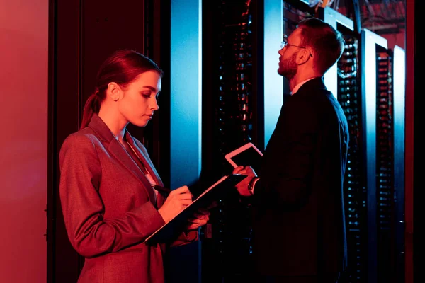 Attractive businesswoman holding clipboard near businessman with digital tablet in data center — Stock Photo