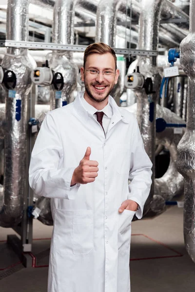 Ingeniero feliz en gafas y abrigo blanco de pie con la mano en el bolsillo y mostrando el pulgar hacia arriba - foto de stock