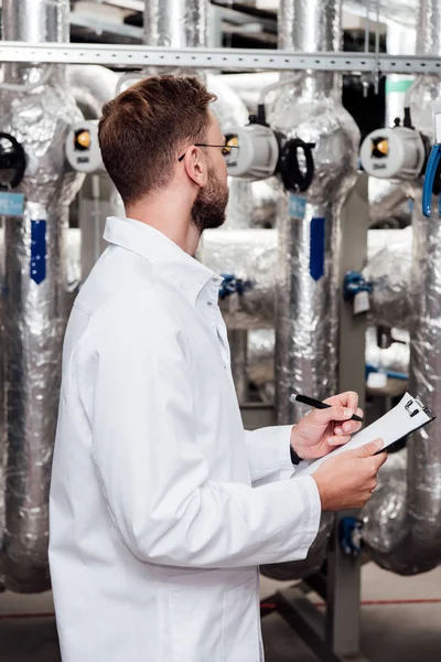 Bearded engineer in white coat holding clipboard and pen near air compressed system — Stock Photo