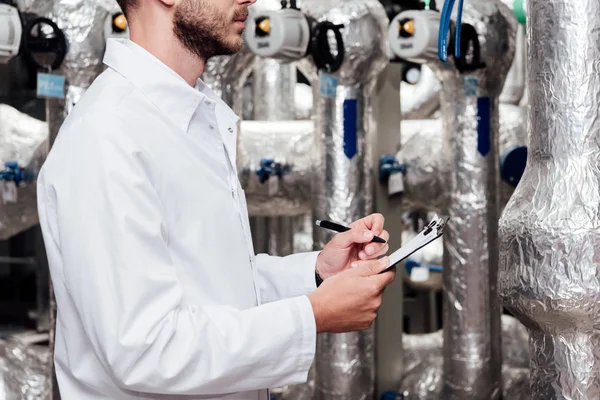 Ausgeschnittene Ansicht eines bärtigen Ingenieurs in weißem Mantel, der Klemmbrett und Stift in der Nähe einer Druckluftanlage hält — Stockfoto