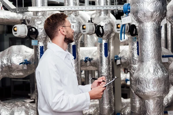 Bearded engineer in white coat holding pen and clipboard near air compressed system — Stock Photo