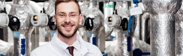 Panoramic shot of happy bearded engineer in white coat near air compressed system — Stock Photo