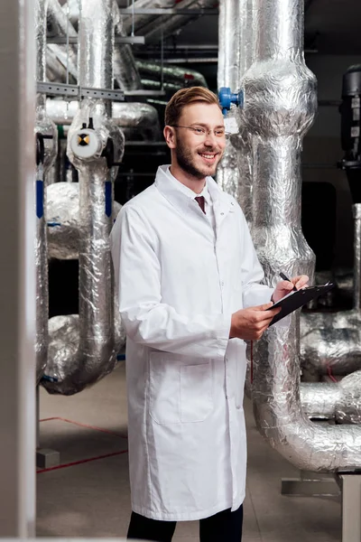Happy engineer in white coat holding pen and clipboard near air compressed system — Stock Photo