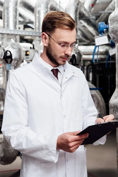 Bearded engineer in white coat holding pen near clipboard and air compressed system — Stock Photo
