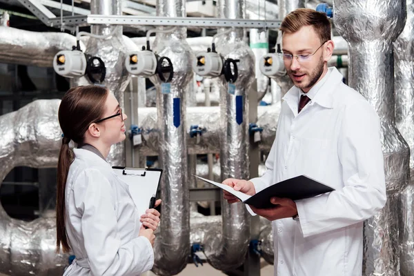 Mujer feliz en blanco abrigo mirando guapo ingeniero con carpeta - foto de stock