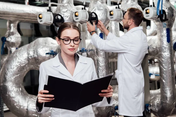 Selective focus of attractive engineer in white coat looking at folder near coworker and compressed air system — Stock Photo